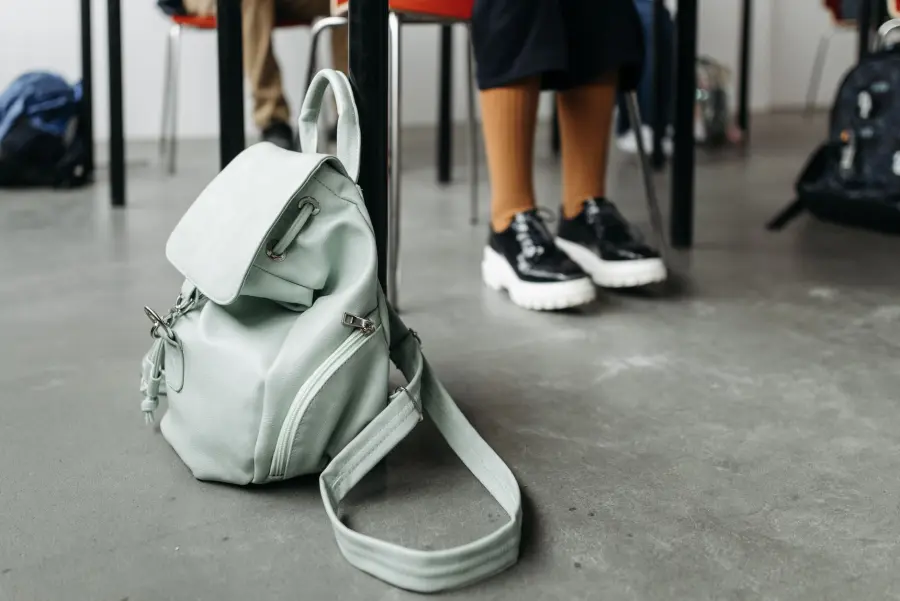 A mint green backpack placed on a concrete classroom floor, partially showing a student's legs.